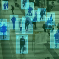 Image of a crowd of people seen through a surveillance camera. Blue boxes appear around the people to represent facial recognition surveillance. The cast of the image is green. 