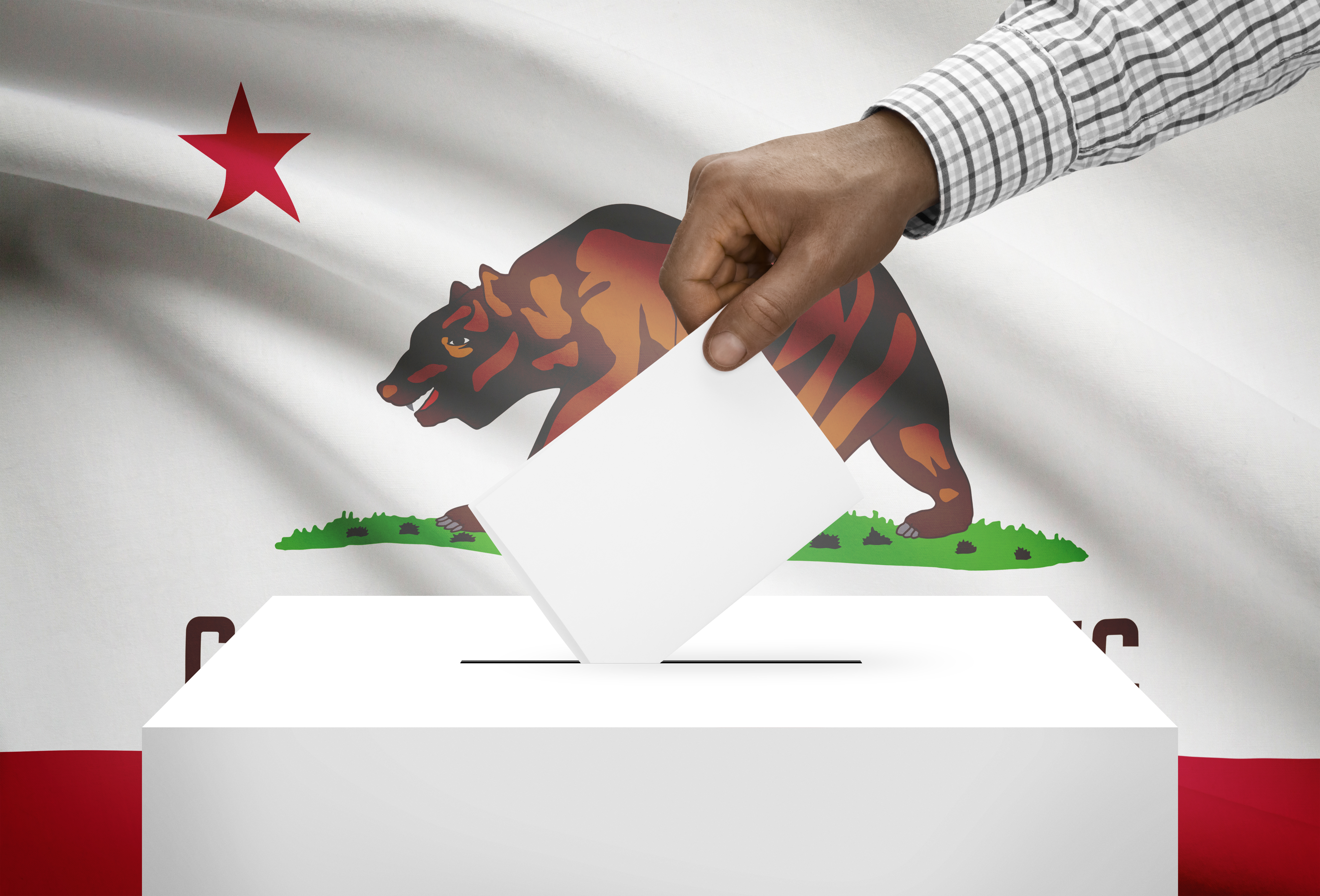 A hand placing a ballot into a ballot box with the California state flag in the background. 