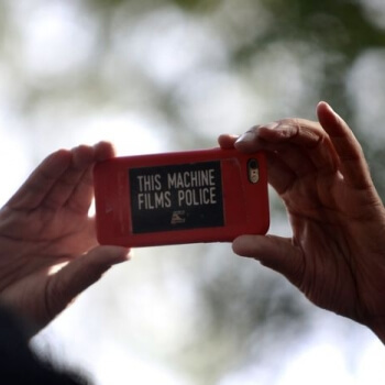 Hands holding a cell phone with a sticker that reads "this machine films police"