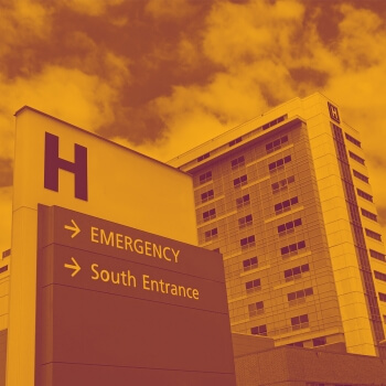 A hospital sign is in the foreground with the building in the background. On the sign there are two options: One says "Emergency" and one says "South Entrance" 