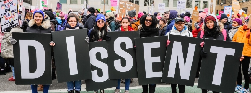 Cropped photograph of seven individuals each holding a letter to spell out 'DISSENT' in front of a group of people who are also holding signs