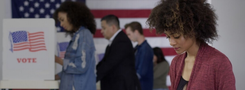 Young woman of color staffing desk at polling station with various voters in background, US flag on wall behind them.