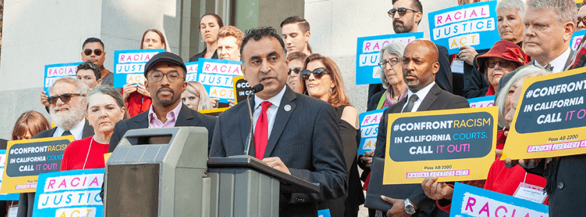 Group of people with racial justice act signs surrounding a speaker on the steps of a capital building