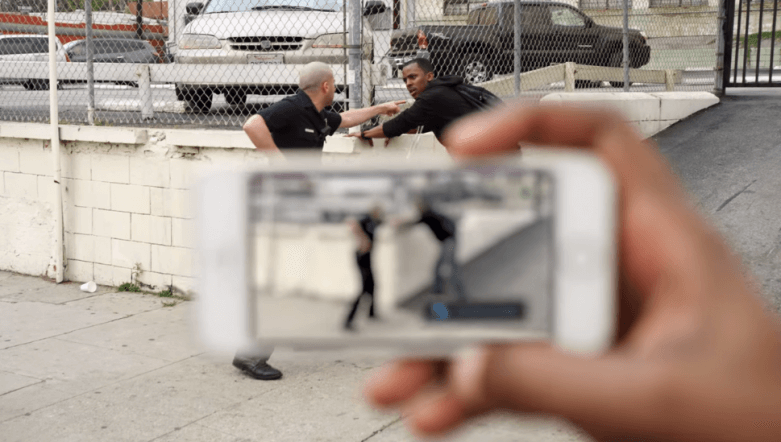 A police officer is gesturing at a man in the background. In the foreground, a hand is holding a phone recording the encounter.