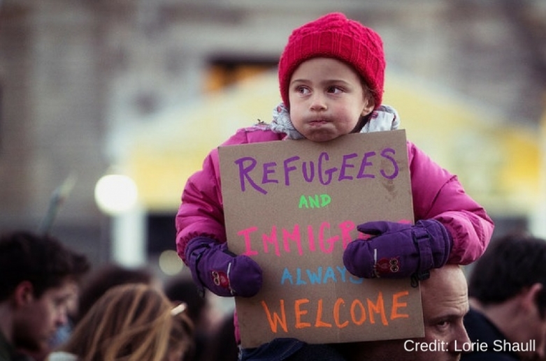 A small child sits on her father's shoulders at a protest, holding a sign that reads "Refugees and immigrants always welcome"