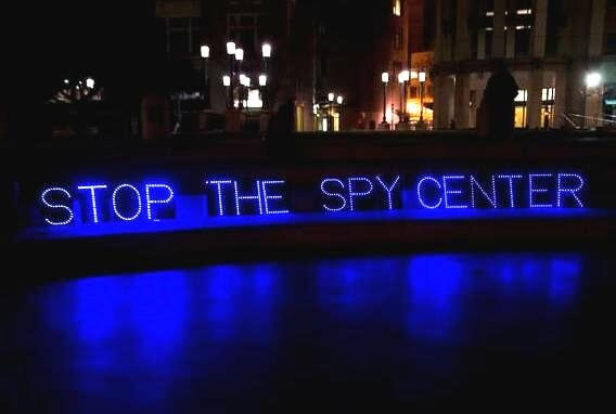 Blue lights in Oakland plaza at night spell out STOP THE SPY CENTER