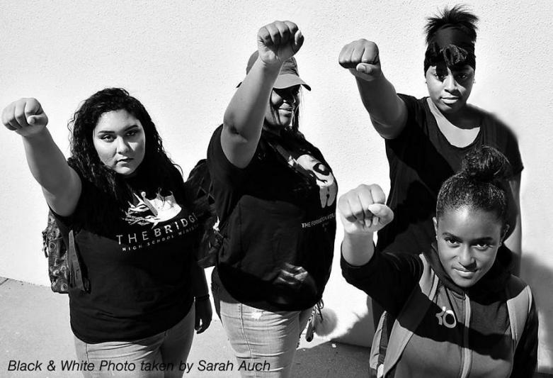 Four female students stand with raised fists, looking up at the camera