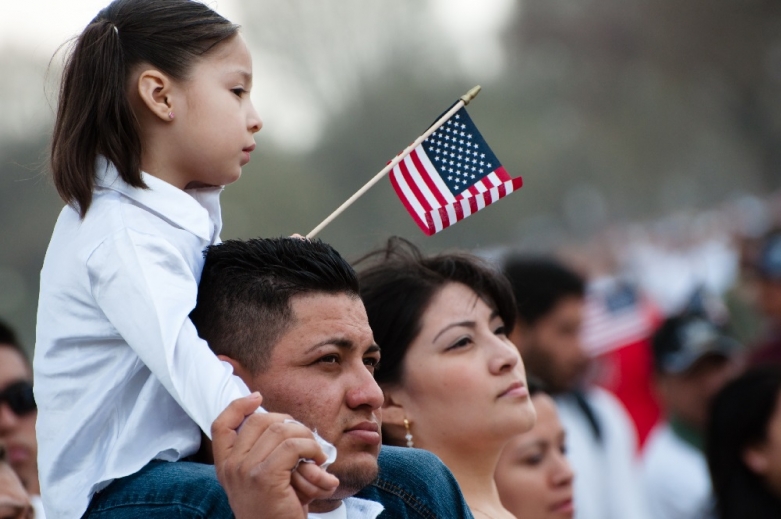 A family at a rally. A girl sits on her father's shoulders, holding an American flag. Her mother stands next to them.