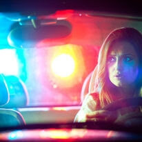 A woman looks in her car's rear-view mirror as police lights flash in the background. She is being pulled over. 