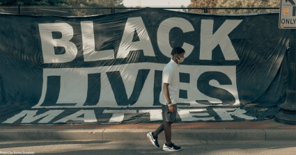 Young black man walking in front of a fence with a large 'Black Lives Matter' banner attached to it.