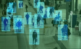 Image of a crowd of people seen through a surveillance camera. Blue boxes appear around the people to represent facial recognition surveillance. The cast of the image is green. 
