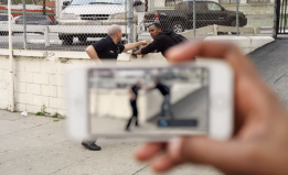 A police officer is gesturing at a man in the background. In the foreground, a hand is holding a phone recording the encounter.