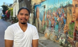 Picture of Jose Ruben. Young, latino man wearing a white t shirt. He is standing next to a colorful mural