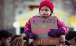 A small child sits on her father's shoulders at a protest, holding a sign that reads "Refugees and immigrants always welcome"
