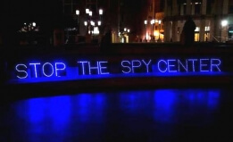 Blue lights in Oakland plaza at night spell out STOP THE SPY CENTER