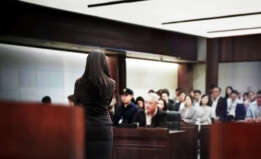 Court room with a person standing and several individuals seated in rows behind observing the proceedings.