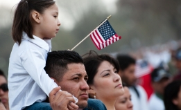 A family at a rally. A girl sits on her father's shoulders, holding an American flag. Her mother stands next to them.