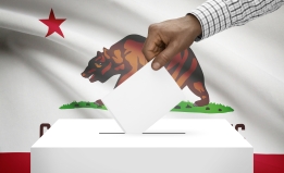 A hand placing a ballot into a ballot box with the California state flag in the background. 