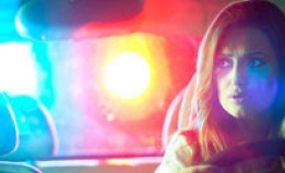 A woman looks in her car's rear-view mirror as police lights flash in the background. She is being pulled over. 